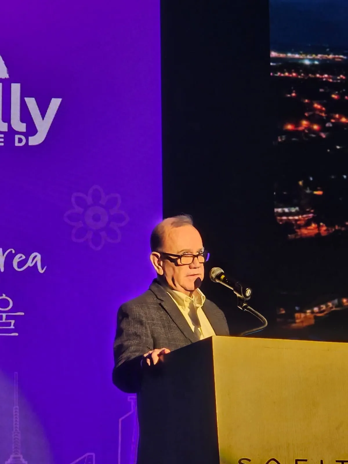 Kiko speaking at a podium in front of a large screen at an event in Seoul